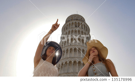 Two attractive women show to each other something on the background of the Leaning Tower. 135178996