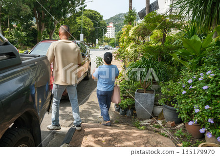 Two people walking along a street with plants and cars under bright sunlight in a city setting 135179970