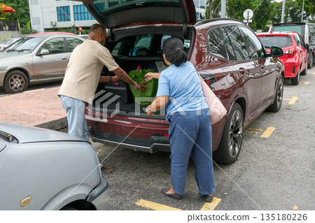 Couple loading groceries into the trunk of a car in a busy parking area on a sunny day Couple loading groceries into the trunk of a car in a busy parking area on a sunny day 135180226