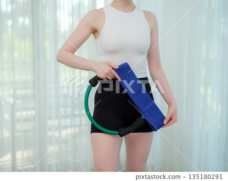 Young woman holding fitness ring and resistance band at home. Concept of home workout, strength training, and healthy lifestyle. Female preparing equipment for online fitness session. 135180291