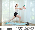 Young woman exercising at home with fitness ring. Concept of strength training, balance, and online workout. Female practicing lower body exercise while following virtual fitness class on tablet. 135180292