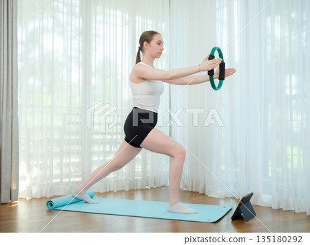 Young woman exercising at home with fitness ring. Concept of strength training, balance, and online workout. Female practicing lower body exercise while following virtual fitness class on tablet. Young woman exercising at home with fitness ring. Concept of strength training, balance, and online workout. Female practicing lower body exercise while following virtual fitness class on tablet. 135180292
