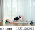 Young woman exercising at home with resistance band on yoga mat. Concept of fitness, healthy lifestyle, and online workout. Female performing glute bridge while following virtual training on tablet. 135180293