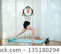 Young woman exercising at home with fitness ring. Concept of strength training, balance, and online workout. Female practicing lower body exercise while following virtual fitness class. 135180294