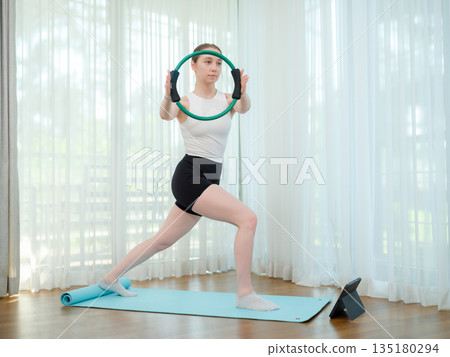 Young woman exercising at home with fitness ring. Concept of strength training, balance, and online workout. Female practicing lower body exercise while following virtual fitness class. 135180294