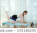 Young woman exercising at home on yoga mat with resistance band. Concept of fitness, strength training, and online workout. Female performing leg raise while following virtual fitness class on tablet. 135180295
