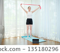 Young woman exercising at home with resistance band. Concept of fitness, healthy lifestyle, and online workout. Female training on yoga mat while following virtual fitness class on a tablet. 135180296