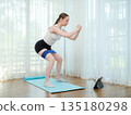 Young woman doing squat exercise at home with resistance band. Concept of fitness, strength, and online workout. Female training on yoga mat while following virtual fitness class on a tablet. 135180298