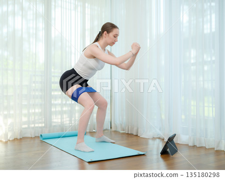 Young woman doing squat exercise at home with resistance band. Concept of fitness, strength, and online workout. Female training on yoga mat while following virtual fitness class on a tablet. 135180298