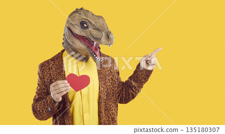 Funny man in dinosaur mask holding red Valentine and pointing at copy space on yellow background 135180307