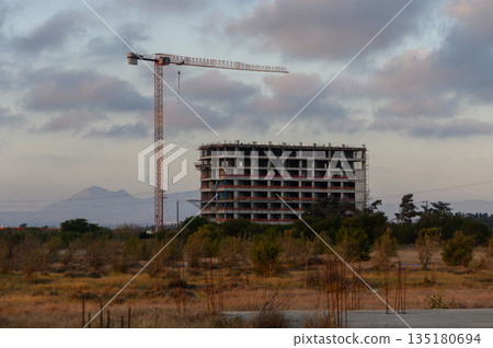 Construction site with towering crane overlooking an unfinished building at sunset in a rural landscape 135180694