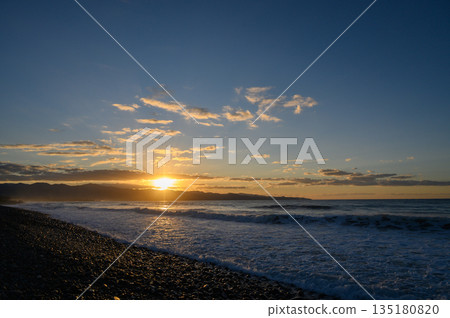 Golden sun setting over a tranquil beach with clouds reflecting vibrant colors on a serene evening 135180820