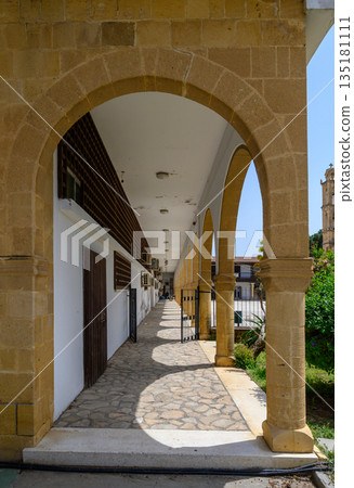 Walking through the historic archway of a charming building in a sunlit village square dotted with greenery 135181111