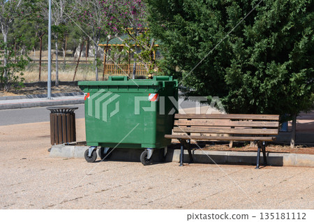 Green dumpster and wooden bench in a serene park setting during a sunny afternoon near blooming trees 135181112