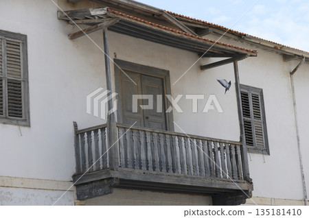 Old wooden balcony with peeling paint and a bird in flight against a clear blue sky 135181410