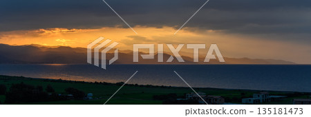 Golden rays break through dark clouds over tranquil sea and distant mountains at dusk Golden rays break through dark clouds over tranquil sea and distant mountains at dusk 135181473