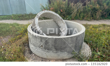 Two concrete well rings of different sizes abandoned in a field near a warehouse. 135182920