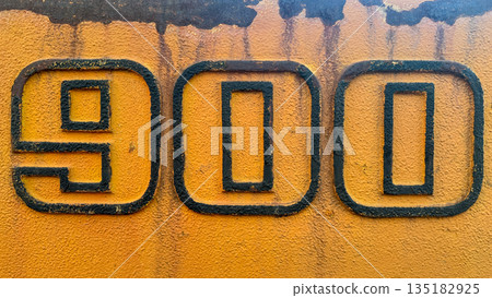 Close-up of industrial number 900 on a weathered yellow textured metal surface. Close-up of industrial number 900 on a weathered yellow textured metal surface. 135182925
