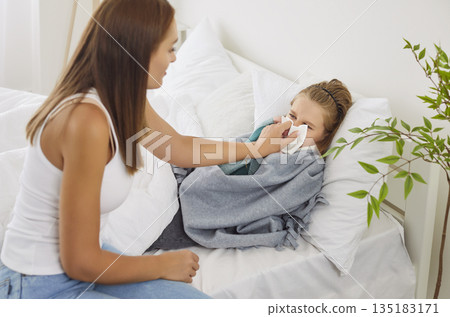 Caring kind young mom and sick child in bed helping to blow nose, sneezing in handkerchief Caring kind young mom and sick child in bed helping to blow nose, sneezing in handkerchief 135183171