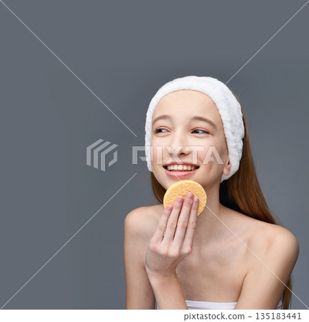 Young caucasian female using facial sponge for skincare routine smiling on grey 135183441
