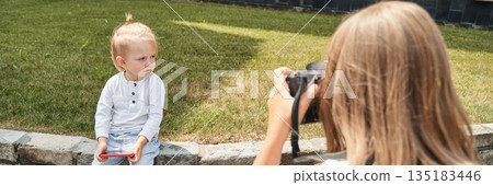 Young child being photographed by woman outdoors with camera in hand Young child being photographed by woman outdoors with camera in hand 135183446
