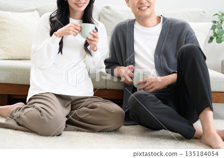 A man and woman relaxing in front of a sofa in the living room with mugs 135184054
