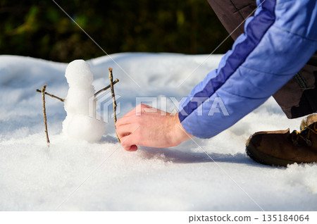 Person Building Tiny Snowman in Fresh Winter Snow With Boots and Blue Jacket 135184064