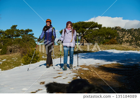 Two Women Hiking With Trekking Poles On Snowy Trail Under Clear Blue Sky Two Women Hiking With Trekking Poles On Snowy Trail Under Clear Blue Sky 135184089