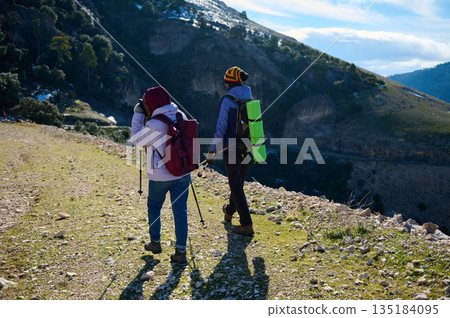 Two Hikers Trek a Mountain Trail With Backpacks And Trekking Poles Under Clear Blue Sky 135184095