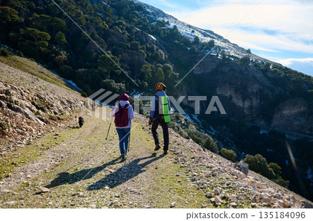 Two Backpackers Hiking Along a Mountain Trail With A Dog Under A Snowy Ridge 135184096