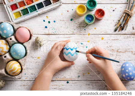 Child hands painting colorful Easter eggs with brush and watercolors on white wooden table background. Child hands painting colorful Easter eggs with brush and watercolors on white wooden table background. 135184246