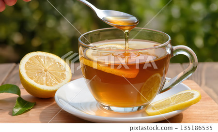 Cup of hot tea with lemon slices and honey being added with spoon on a wooden table outdoors. Natural and healthy drink concept. 135186535