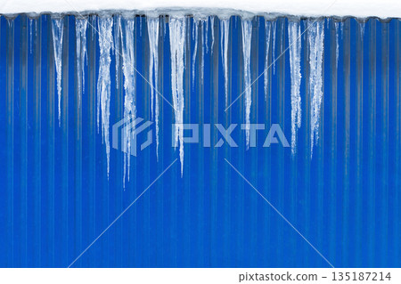 Roof of building with blue color metal wall covered with sharp icicles. Thaw, winter weather 135187214
