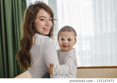 Cozy family moment with a young woman holding her smiling baby at home, standing near soft window light. A warm and cozy parent-child scene filled with love 135187473