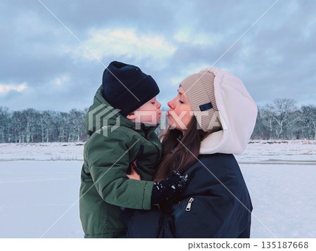 Mother and child sharing tender winter kiss. Loving mom and little boy in warm hats touch noses affectionately on snowy landscape with forest background. 135187668