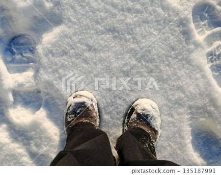 Winter boots standing in fresh snow. POV shot of snowy winter boots with footprints around on bright white snow surface under sunlight. 135187991