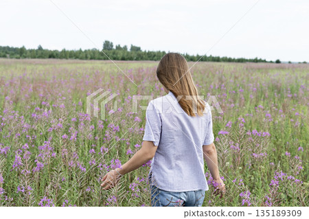 Young beautiful blond woman in purple shirt from behind walking in the meadow among flowers of fireweed 135189309