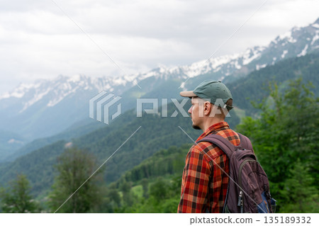 Side view of man with backpack standing on mountain viewpoint , looking at scenic alpine landscape with forested hills and snow-capped peaks in distance Concept of hiking, adventure travel copy space Side view of man with backpack standing on mountain viewpoint , looking at scenic alpine landscape with forested hills and snow-capped peaks in distance Concept of hiking, adventure travel copy space 135189332