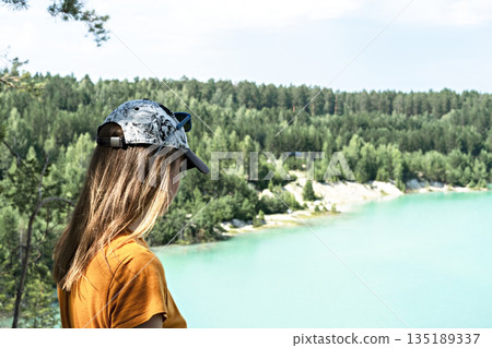Rear view of young blond woman traveler in a cap and mustard t-shirt standing on the cliff of a blue turquoise lake or river and coniferous forest, travel beautiful landscape 135189337