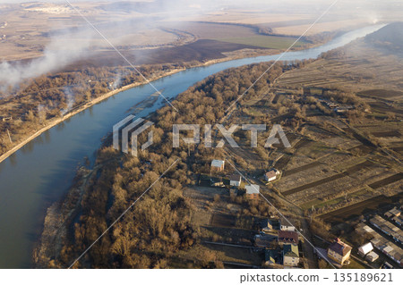 Aerial top view of river flowing through town. Rural landscape of residential house roofs, roads and tree tops on spring or autumn day. Drone photography. Aerial top view of river flowing through town. Rural landscape of residential house roofs, roads and tree tops on spring or autumn day. Drone photography. 135189621