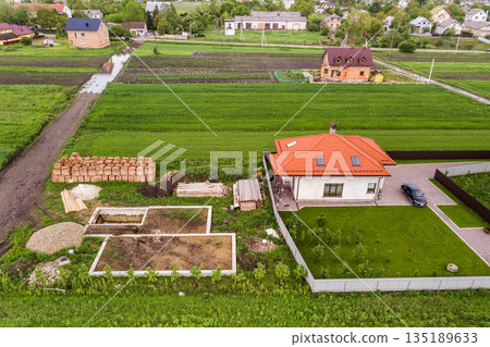 Aerial top view of a private house with attic windows on roof, paved yard with green grass lawn and building site with concrete foundation floor and stacks of yellow bricks for construction.. Aerial top view of a private house with attic windows on roof, paved yard with green grass lawn and building site with concrete foundation floor and stacks of yellow bricks for construction.. 135189633