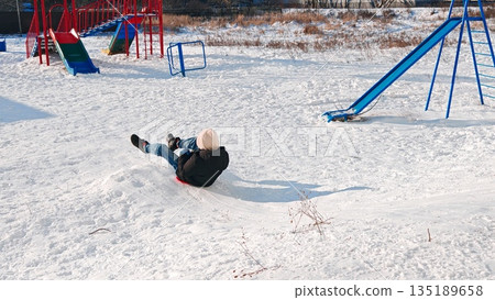 Child slides down snow mound. Child slides on red sled down snow mound at colorful playground. Child slides down snow mound. Child slides on red sled down snow mound at colorful playground. 135189658