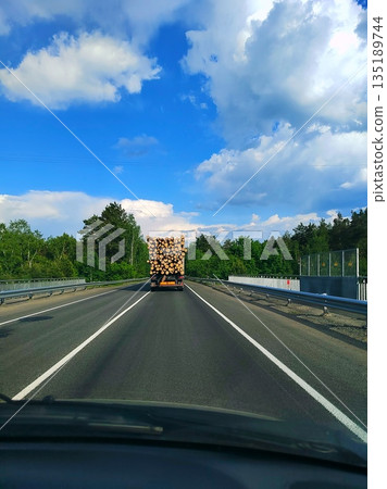 Logging truck carrying timber on highway through forest. Heavy truck loaded with cut logs driving on rural road under blue sky Logging truck carrying timber on highway through forest. Heavy truck loaded with cut logs driving on rural road under blue sky 135189744