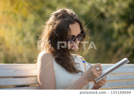 Young woman writing notes in notebook sitting on bench in park 135189899