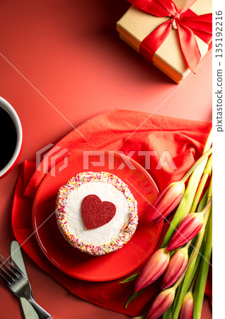 Valentine's Day festive red background. Cake with red heart, cup of coffee, gift box and spring red tulip flower bouquet. Top view Valentine's Day festive red background. Cake with red heart, cup of coffee, gift box and spring red tulip flower bouquet. Top view 135192216