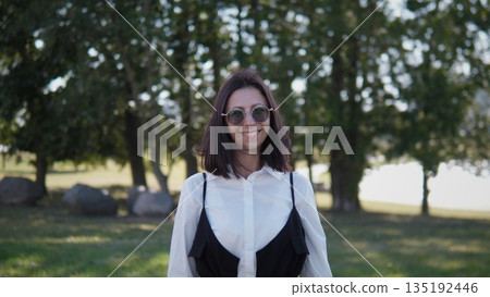A confident woman stands outdoors in a park, wearing sunglasses and a stylish outfit. She smiles while surrounded by trees and natural beauty, enjoying a sunny day. 135192446