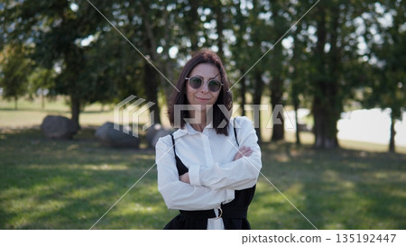 A woman stands confidently in a park, smiling with arms crossed. She wears stylish sunglasses and a black-and-white outfit, enjoying a pleasant day outdoors in a natural setting. 135192447