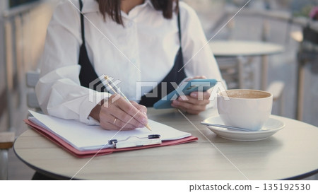 A professional woman sits at a cafe, writing notes on a clipboard while checking her phone. She enjoys a cup of coffee in a relaxed outdoor setting, focused on her tasks. A professional woman sits at a cafe, writing notes on a clipboard while checking her phone. She enjoys a cup of coffee in a relaxed outdoor setting, focused on her tasks. 135192530