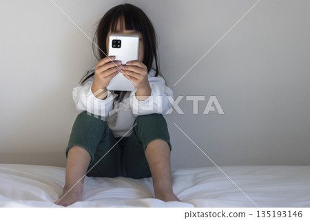 Image of a girl sitting in a dimly lit room looking at her smartphone (5 years old, Japanese, no face) 135193146
