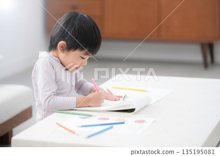 A young child is engrossed in drawing and coloring with colored pencils in a white living room, fostering imagination and creativity. A young child is engrossed in drawing and coloring with colored pencils in a white living room, fostering imagination and creativity. 135195081